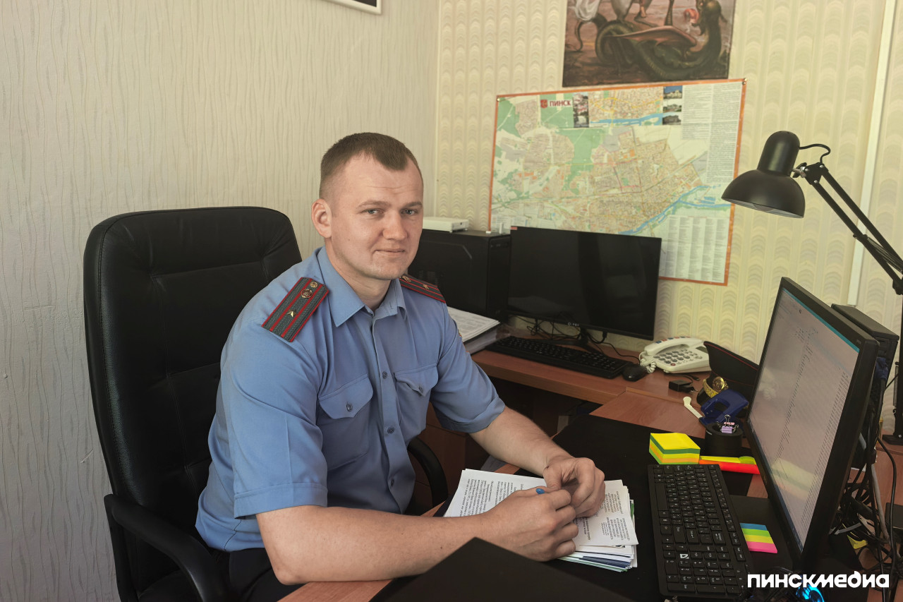 Начальник ИДН Пинского ГОВД в голубой форменной рубашке работает за компьютером на фоне карты города в служебном кабинете.