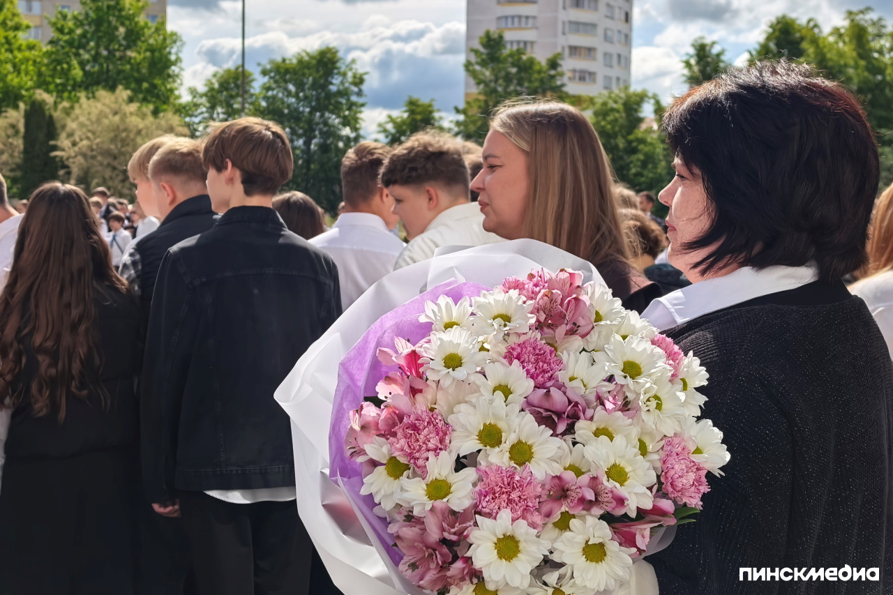 Выпускники во дворе школы и учитель с цветами.